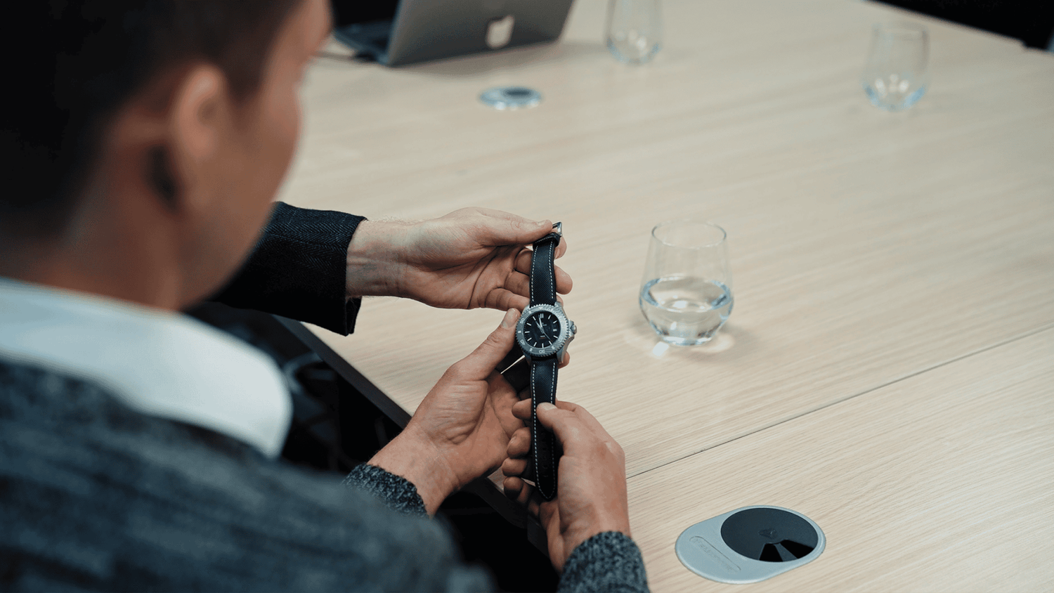 Person adjusting a Damascus Steel watch in an office setting with a glass of water on a table.