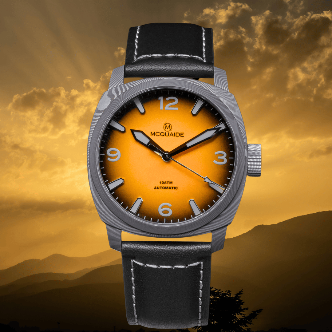 McQuaide automatic wristwatch with a silver-tone case, black leather strap, and a bright orange dial, set against a golden sunset mountain background.