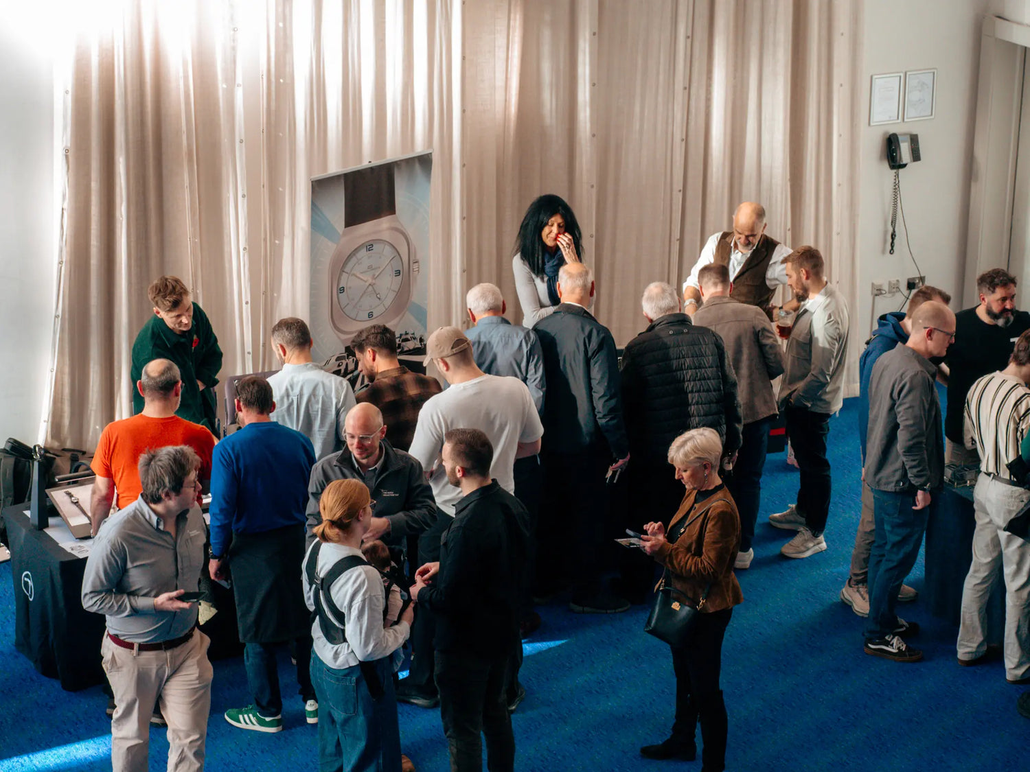 Group of people gathered in a room with a large poster displaying a watch